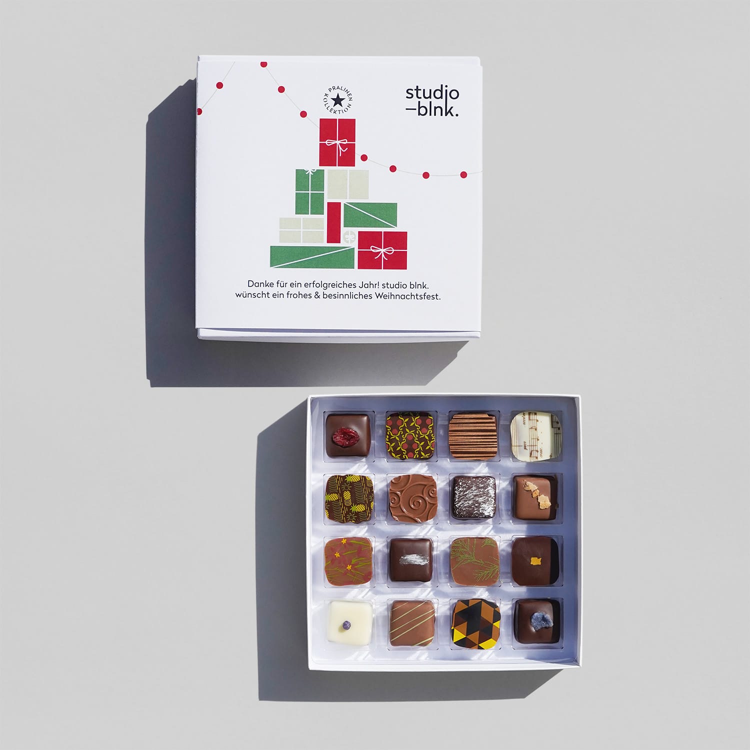 Zwei quadratische weiße Pralinen-Boxen, eine geöffnet mit 16 bunten Pralinen. Verschieden dekoriert mit farbigen Mustern oder Nüssen.
Das weihnachtliche Motiv auf der Pralinenbox ist ein Stapel mit Geschenken in Form eines Tannenbaums und einer Lichterkette. Die Farben sind rot, grün und schwarz.