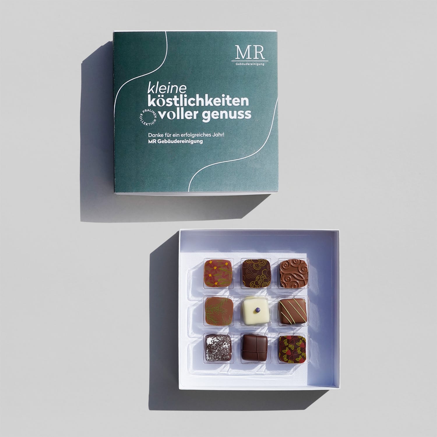 Firmenkunden_Muster-Pralinen_9_MR Zwei quadratische weiße Pralinen-Boxen, eine geöffnet mit 9 bunten Pralinen. Verschieden dekoriert mit farbigen Mustern oder Nüssen.
Die Banderole um die Box ist dunkelgrün mit einem Verlauf, es gibt weiße Linien und einen weißen Text: Kleine Köstlichkeiten voller Genuss.
Es gibt ein Muster für Text und Logo. Diese können Firmenkunden selbst einsetzen.