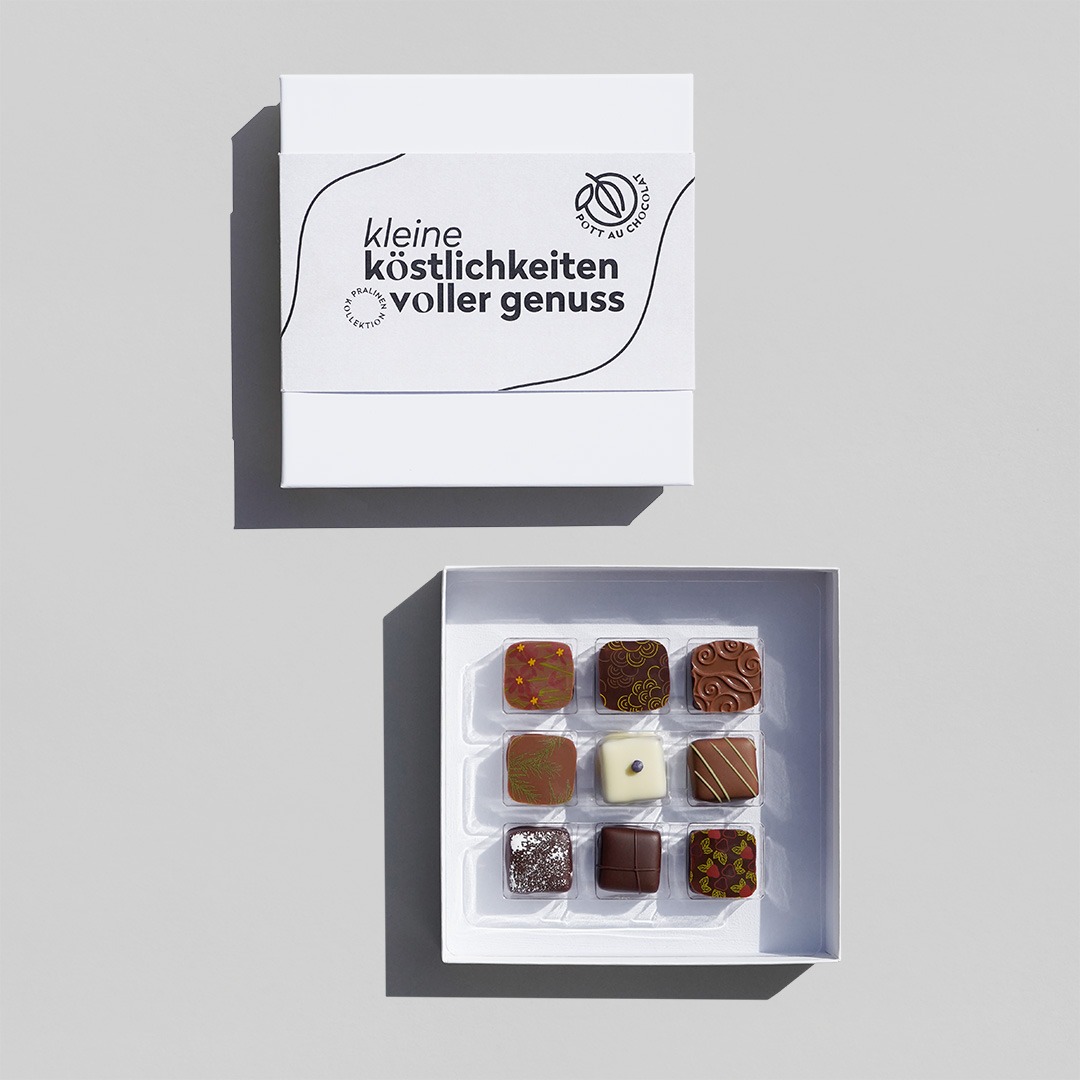 Geöffnete Pralinenschachtel mit 9 bunt dekorierten Pralinen in einer weißen Schachtel. Darüber liegt die geschlossene Schachtel mit einer schmalen Banderole mit der Aufschrift 'kleine Köstlichkeiten voller Genuss' in grau / schwarz.