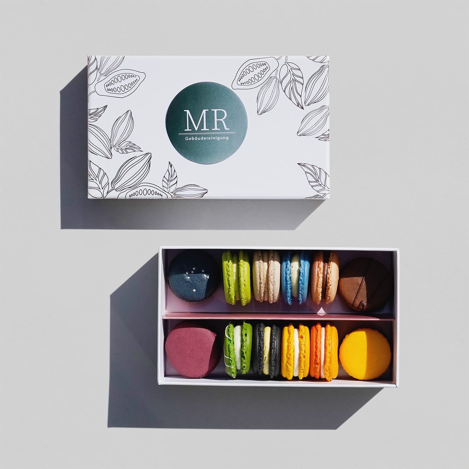 Eine geöffnete weiße Box mit 12 bunten Macarons. Daneben der passende Deckel mit geprägtem Muster aus Kakaofrüchten in Kupfer. Darauf ein dunkelgrüner Sticker mit Verlauf und weißem Muster-Logo.