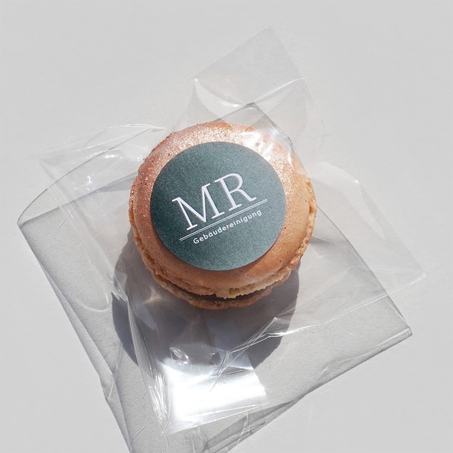 Ein Macaron in einer transparenten Plastik Verackung. Darauf ein dunkelgrüner Sticker mit Verlauf und weißem Muster-Logo.