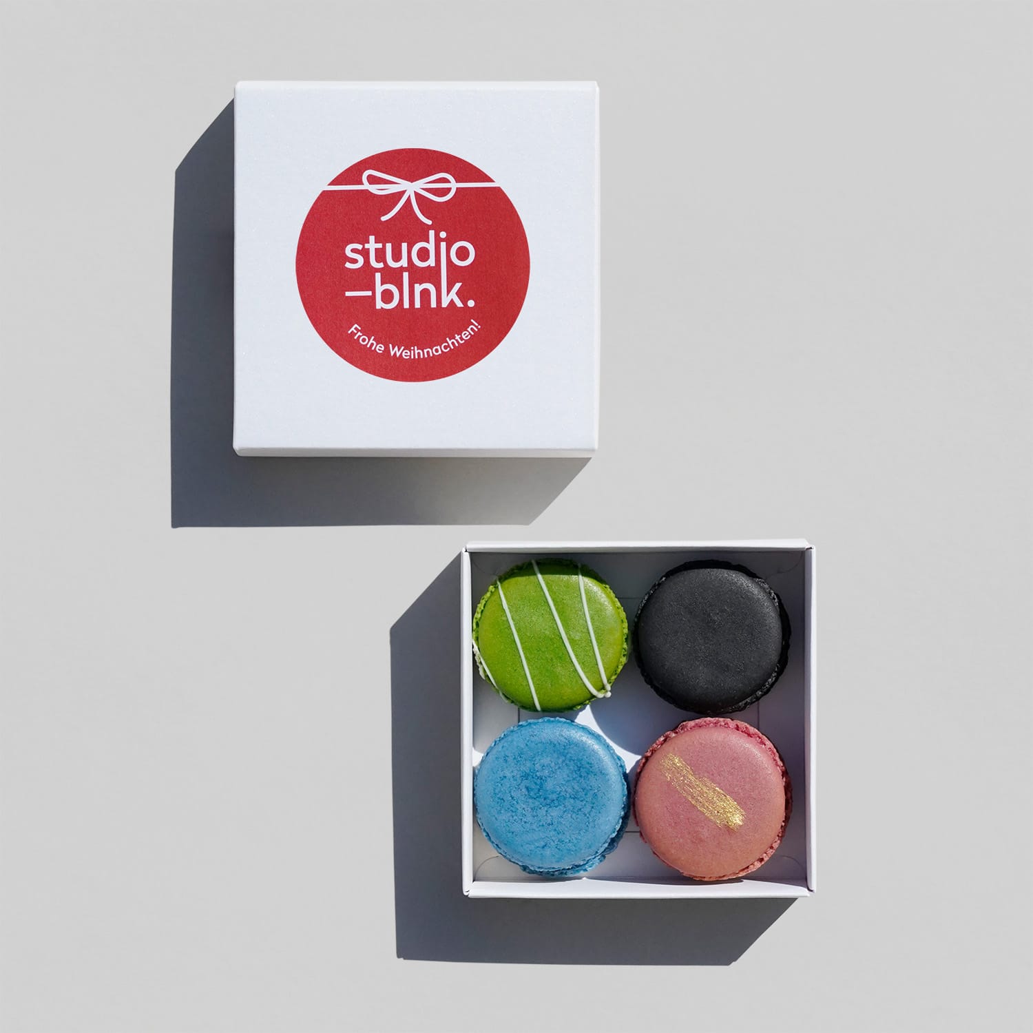 Eine geöffnete weiße Box mit 4 bunten Macarons. Daneben der passende Deckel mit Sticker in rot mit weißem Muster-Logo und 'Frohe Weihnachten'
