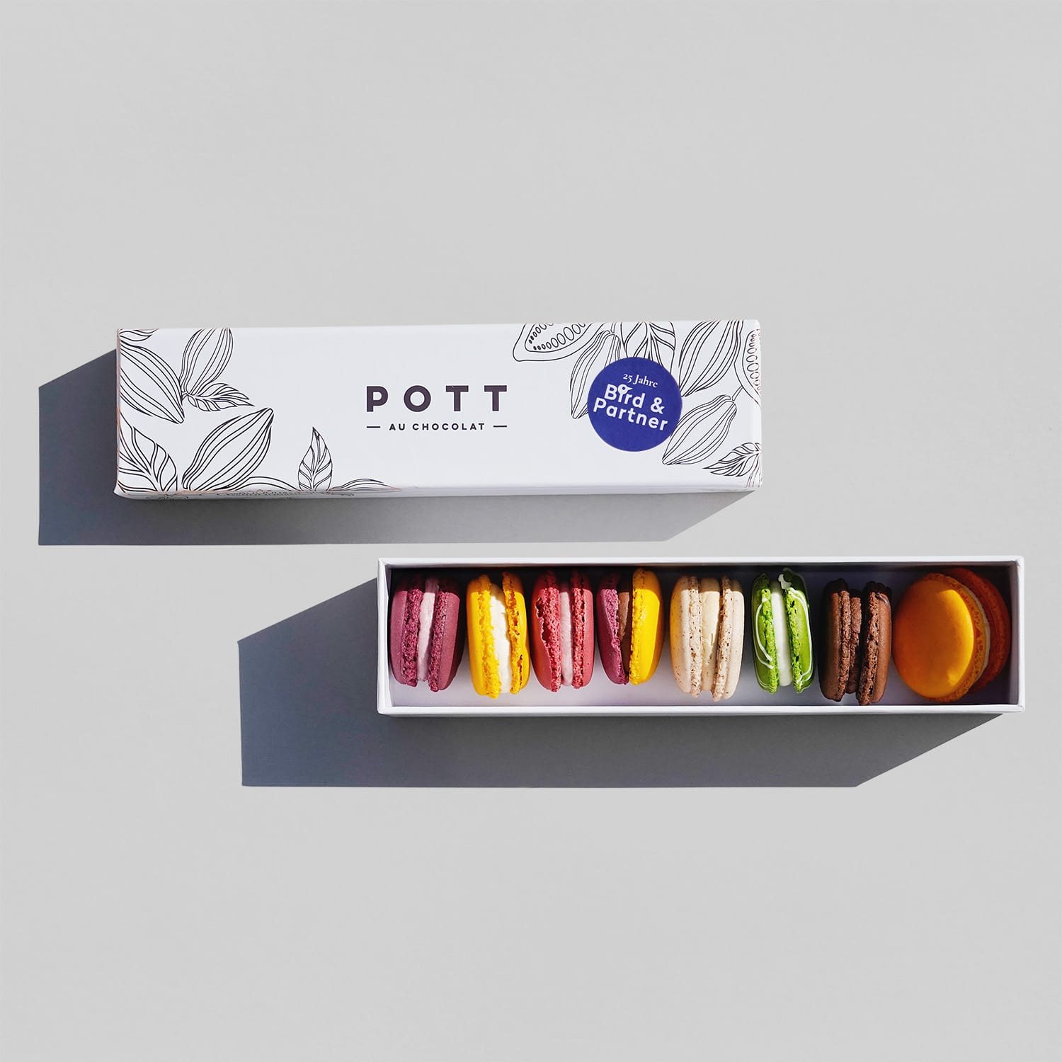 Eine geöffnete weiße Box mit 8 bunten Macarons. Daneben der passende Deckel mit geprägtem Muster aus Kakaofrüchten in Kupfer. Darauf ein blauer Sticker mit weißem Muster-Logo.