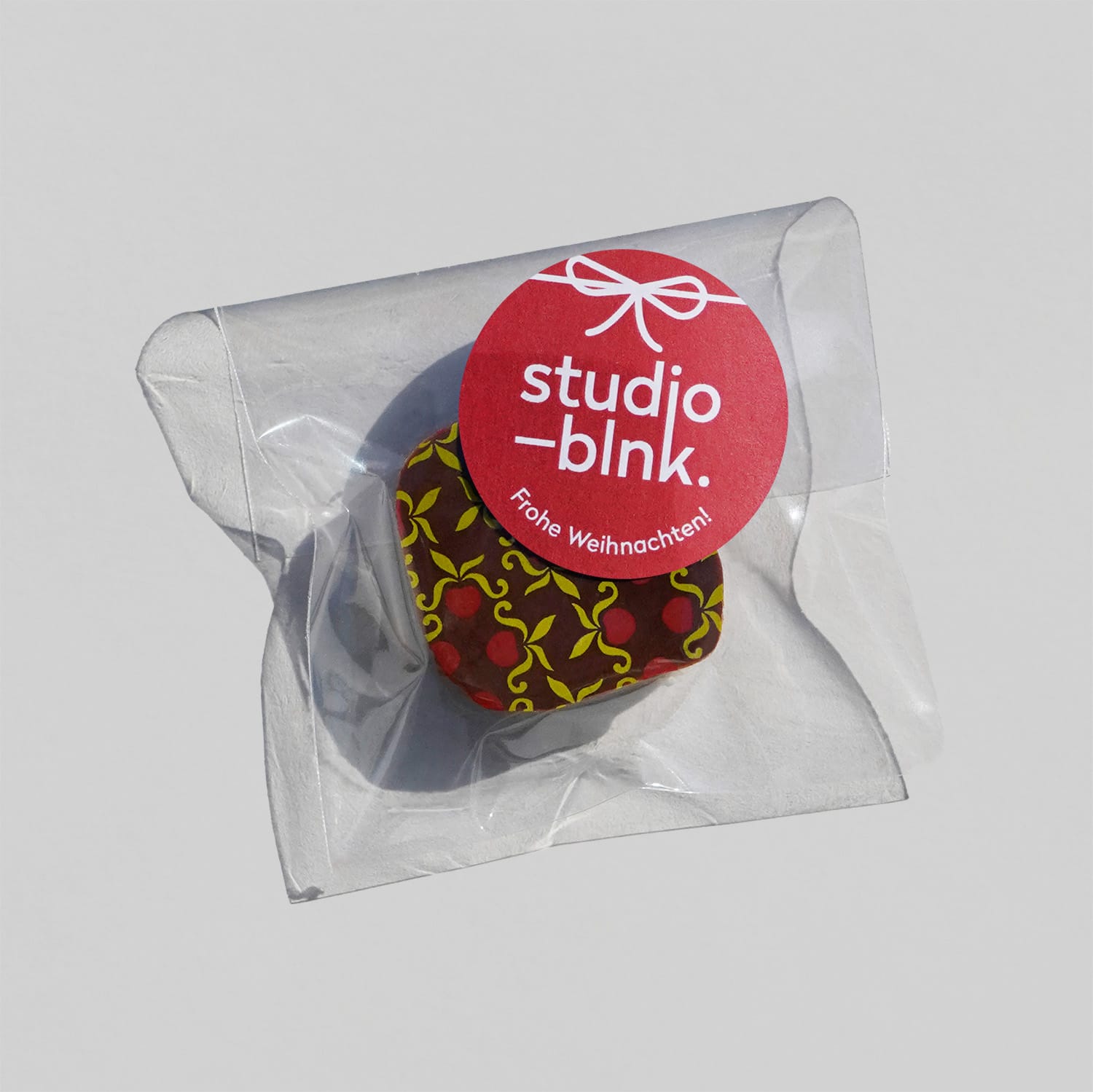 Eine Praline mit dunkler Schokolade und grün rotem Muster in einem transparenten Tütchen. Darauf ist ein runder Sticker in rot mit einem Musterlogo in weiß, einer Schleife und 'Frohe Weihnachten'