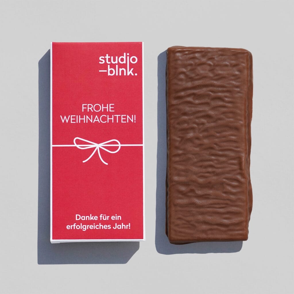 Eine Milchschokoladen-Tafel. Daneben die passende Verpackung aus Pappe rot bedruckt mit einem Musterlogo in weiß, einer Schleife und 'Frohe Weihnachten'
