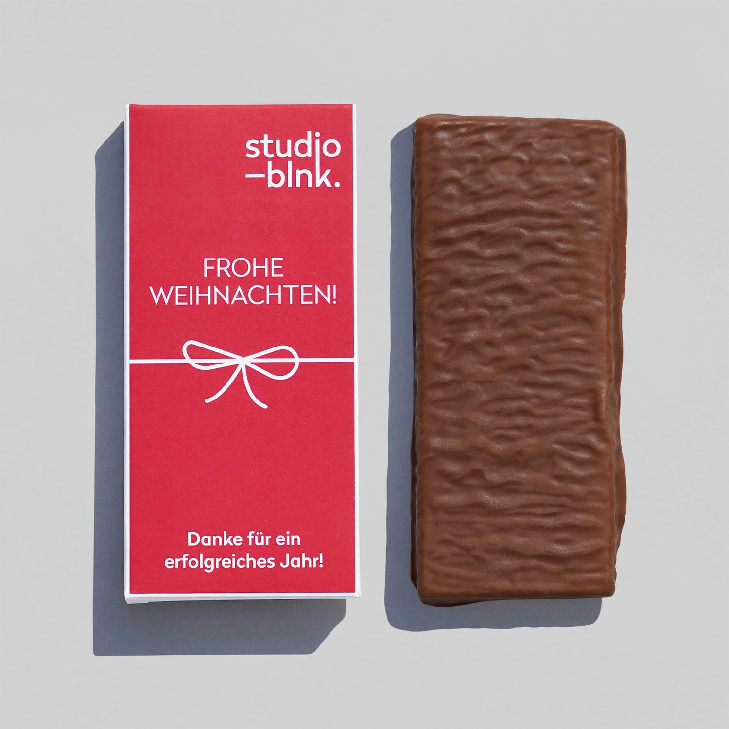 Eine Milchschokoladen-Tafel. Daneben die passende Verpackung aus Pappe rot bedruckt mit einem Musterlogo in weiß, einer Schleife und 'Frohe Weihnachten'