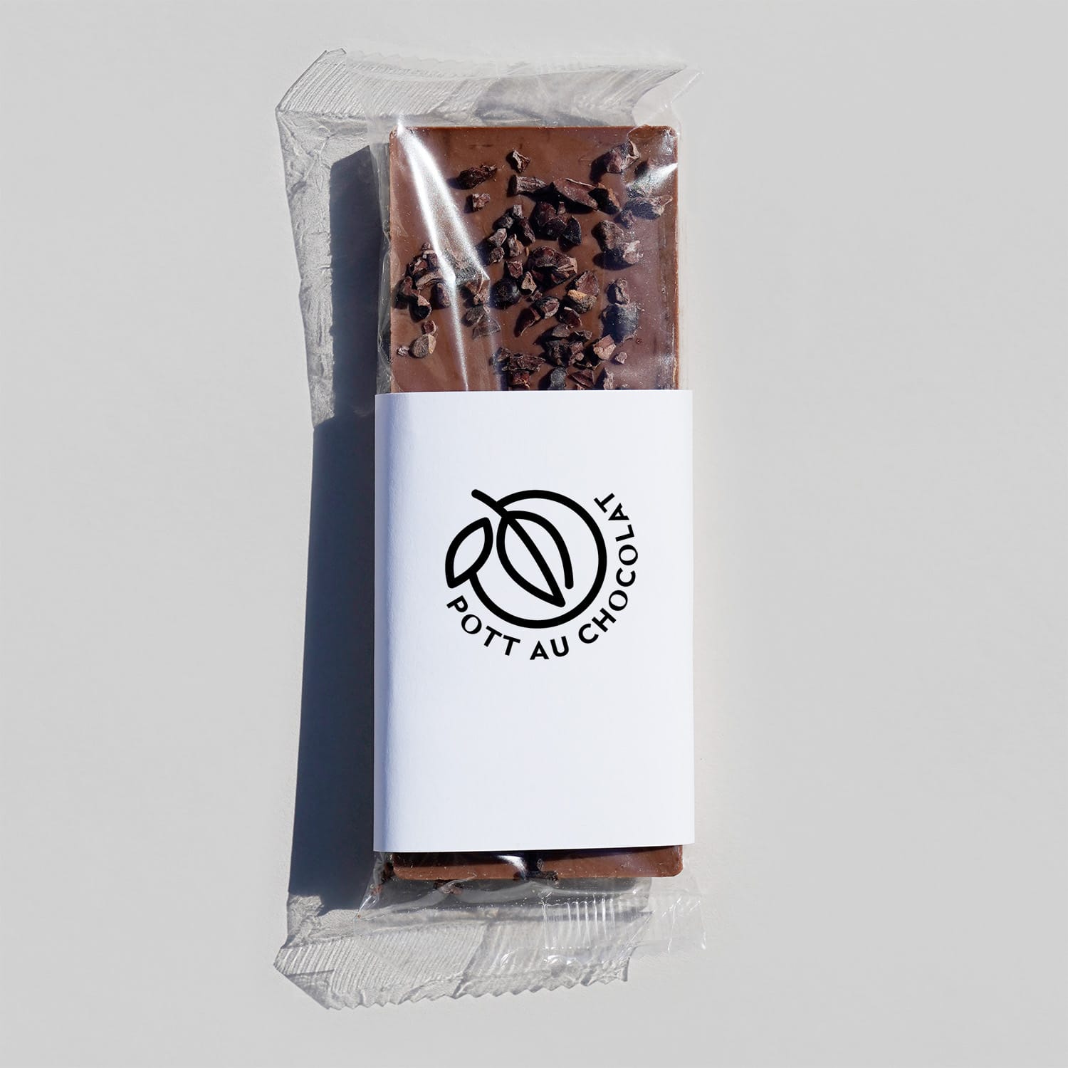Schokoladentafel belegt mit Kakaosplittern in einer transparenten Plastik Verpackung mit einer weißen Banderole, auf der das Pott au Chocolat Logo in schwarz gedruckt ist.