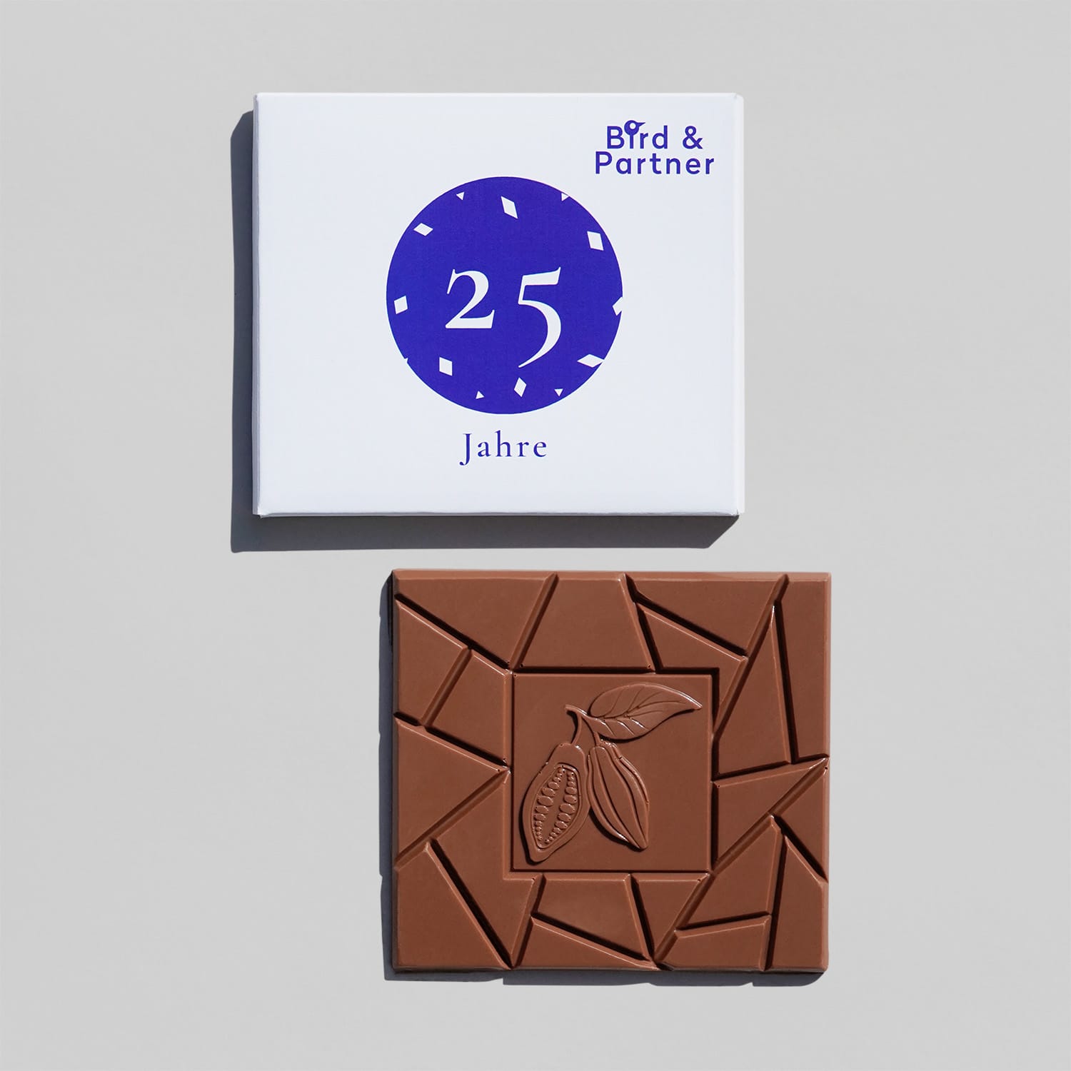 Eine quadratische Milchschokoladen Tafel und die passende Verpackung dazu.
Die Verpackung aus Pappe ist weiß mit einem Motiv mit blauem Kreis, mit weißem Konfetti und '25 Jahre' und ein Muster Logo.