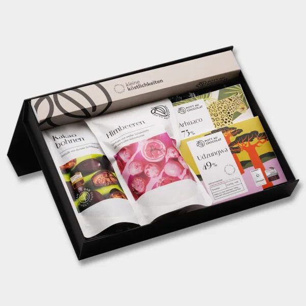 Geschenkbox mit verschiedenen Produkten von Pott au Chocolat: Zwei Feinkosttüten – Himbeeren umhüllt von weißer Schokolade und schokolierte Kakaobohnen. Zusätzlich eine Pralinenstange mit 10 Pralinen und zwei Tafeln Schokolade."