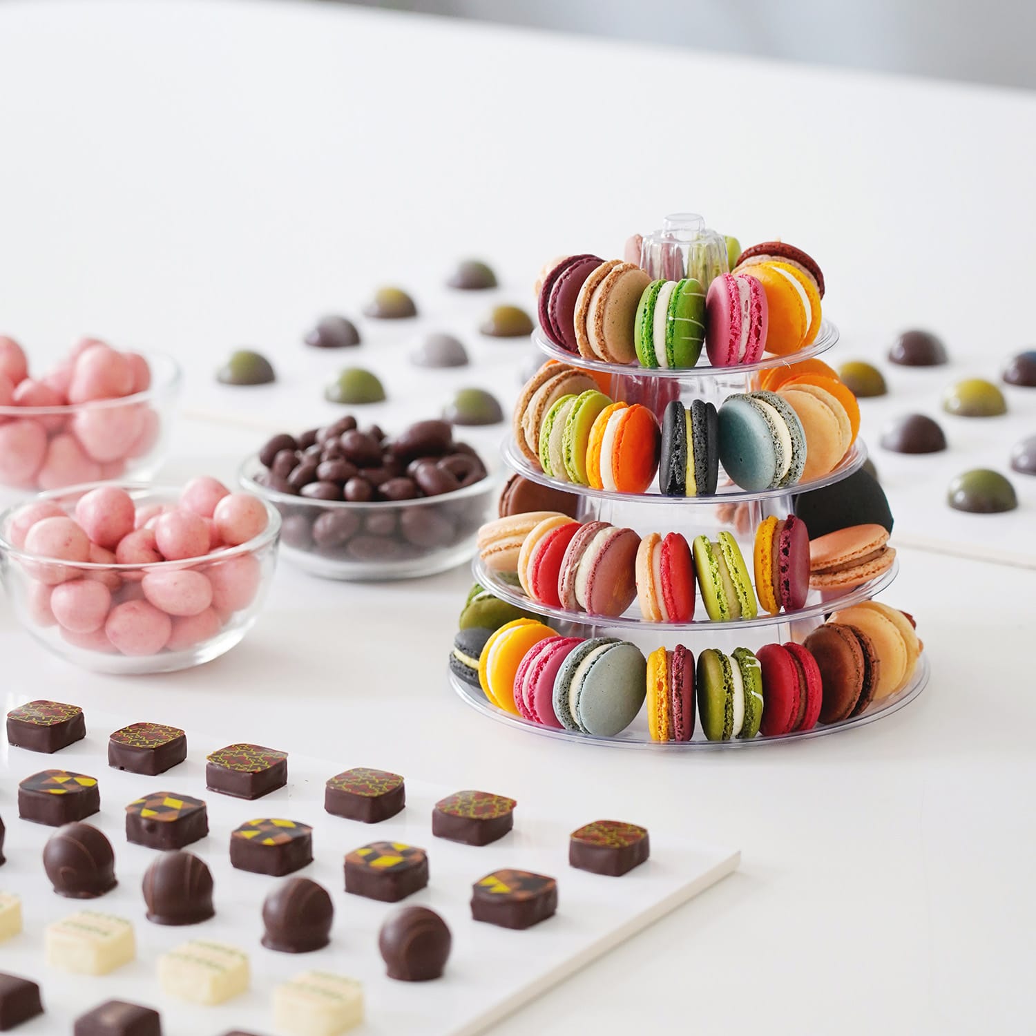 Ein kleines Buffet mit Schokoladenprodukten ist auf einem weißen Tisch aufgebaut. Es gibt zwei Platten mit verschiedenen bunt dekorierten Pralinen, Schälchen mit Nüssen und Früchten, die von Schokolade umhüllt sind und eine Art Pyramide mit ca. 30 sichtbaren, bunten Macarons 