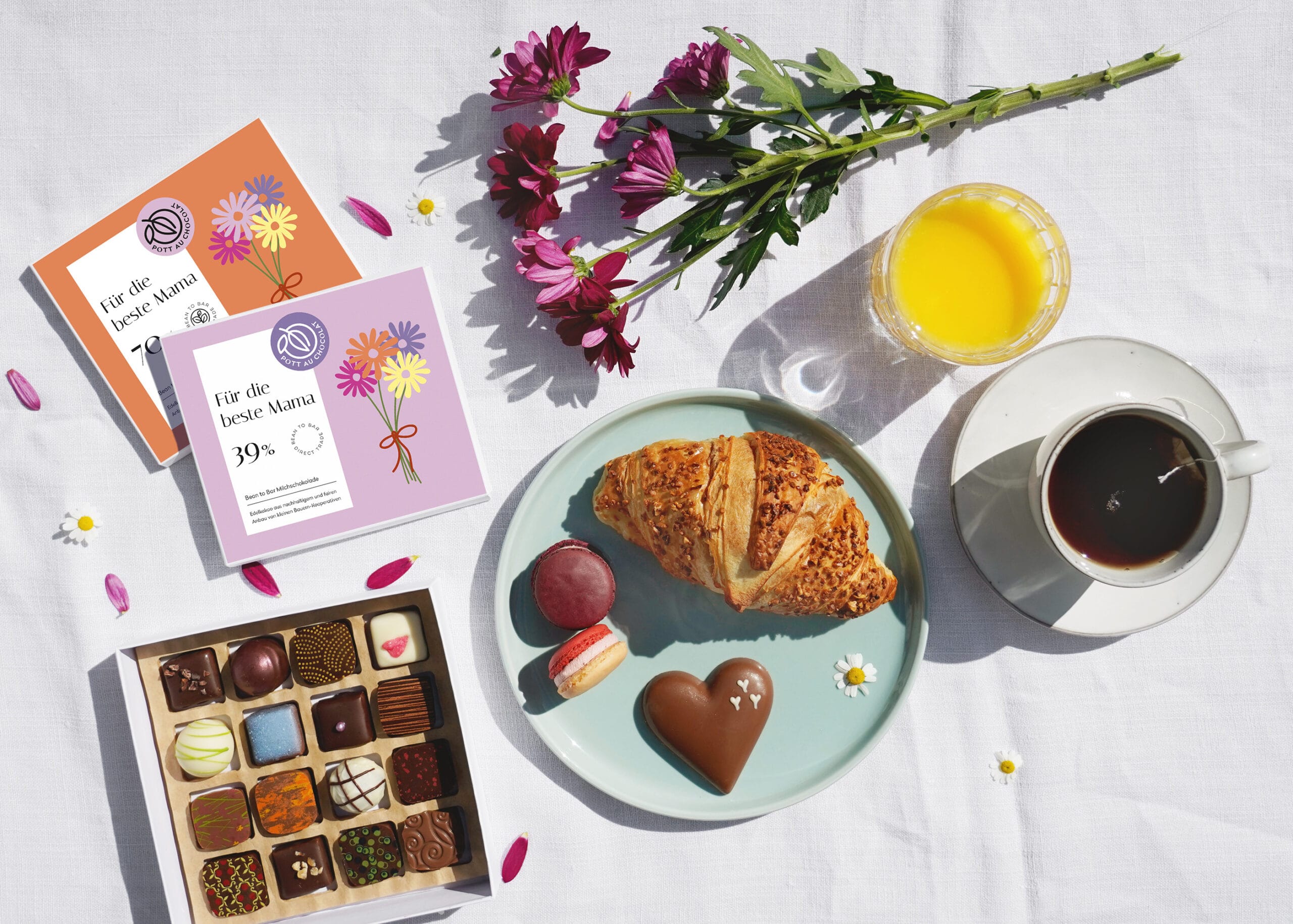 Frühstück zum Muttertag. Zwei Schokoladentafeln mit Blumenmotiv und 'Für die beste Mama', eine geöffnete Pralinenschachtel mit 16 bunten Pralinen, Blumenstrauß, Croissiant, Teetasse und Orangensaft zum Muttertag.