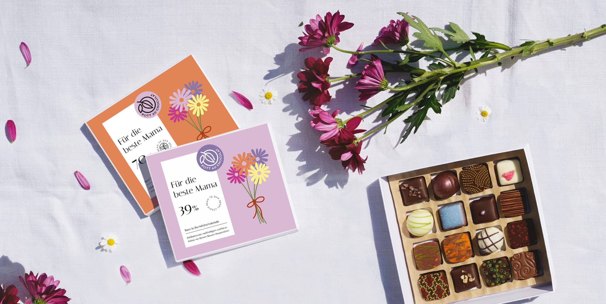 Von oben fotografiert sieht man verschiedene Dinge auf einer weißen Tischdecke liegen. Zwei Schokoladentafeln in ihrer Verpackung in orange und rosa, jeweils mit einer Illustration, die einen bunten Blumenstrauß zeigt. Man kann die Aufschrift 'Für die beste Mama' lesen. Außerdem sind auf dem Foto noch ein Blumenstrauß zu sehen, sowie eine geöffnete Pralinen Schachtel mit 16 Pralinen, teilweise bunt dekoriert mit dunkler, weißer und Milchschokolade.