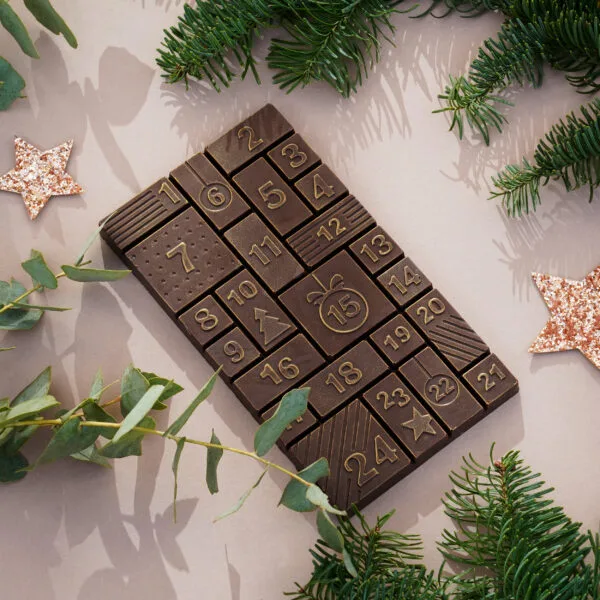 Schokoladentafel in dunkler Schokolade in Adventskalenderform gegossen. Die einzelnen Stückchen der Schokolade zeigen die Zahlen 1-24. Die Schokolade glänzt leicht gold und liegt auf beigem Hintergrund. Daneben verschiedene Zweige und Sterne.