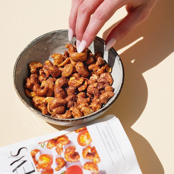 Eine Hand greift in eine Schüssel, die gefüllt ist mit Cashewkernen. Daneben liegt im Anschnitt die Verpackung.