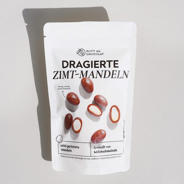 Verpackung der dragierten Zimt-Mandeln. Weiße PET Verpackung mit Aufdruck. Man sieht die Dragierten Mandeln, teilweise aufgeschnitten, und mit Milchschokolade umhüllt.