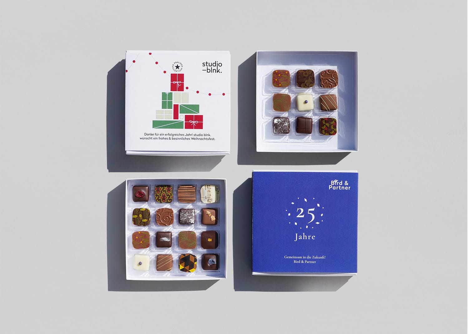 Zwei Pralinenschachteln geöffnet mit jeweils 9 bzw. 16 Pralinen. Sie sind bunt dekoriert und in weißer, dunkler und Milchschokolade. Die Verpackungen sind mit Designs zu Weihnachten und einem Firmenjubiläum bedruckt.