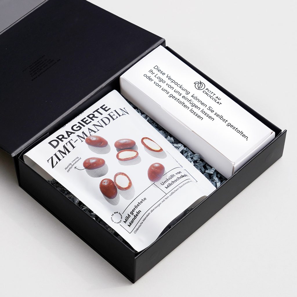 Eine geöffnete schwarze Geschekbox mit einer Tüte mit dragierten Zimt Mandeln und einer weißen Schachtel für 10 Pralinen, die selbst gestaltet werden kann.