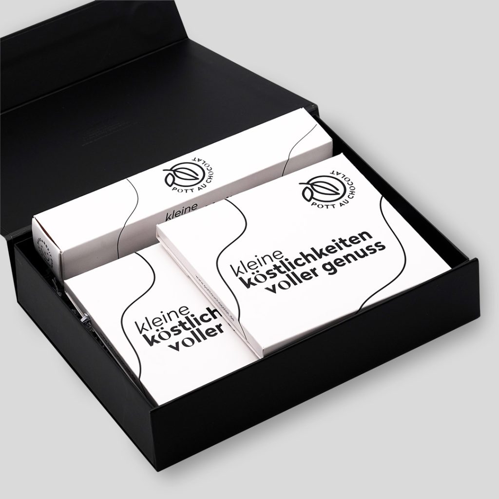 Schwarze Geschenkbox geöffnet und gefüllt mit einer Pralinen Stange für 6 Pralinen und zwei Tafeln von Pott au Chocolat. Die Produkte sind weiß mit schwarzen Illustrationen und Logo von Pott au Chocolat. Sie können von allen Seiten mit Wunschdesign bedruckt werden.