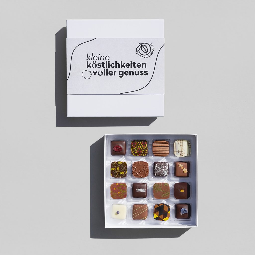 Geöffnete Pralinenschachtel mit 16 bunt dekorierten Pralinen in einer weißen Schachtel. Darüber liegt die geschlossene Schachtel mit einer schmalen Banderole mit der Aufschrift 'kleine Köstlichkeiten voller Genuss' in grau / schwarz.