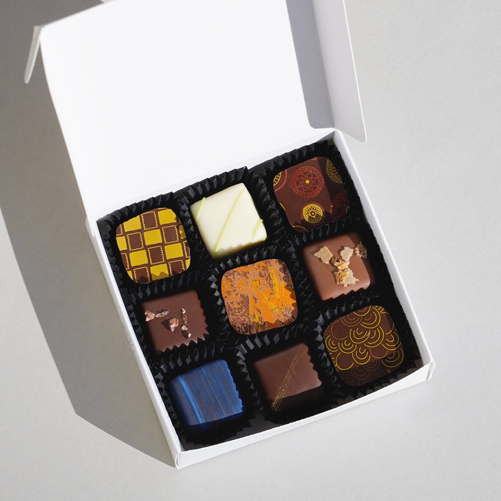 Weiße Pralinenschachtel mit geöffnetem Deckel und 9 bunt dekorierten Pralinen in schwarzen Kapseln.