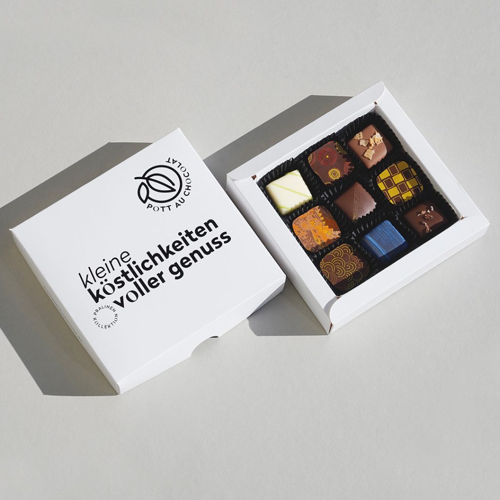 Weiße Pralinenschachtel mit der Aufschrift 'Kleine Köstlichkeiten voller Genuss'. Die Box ist geöffnet, der Deckel liegt separat. In der Box befinden sich 9 bunt dekorierte Pralinen.