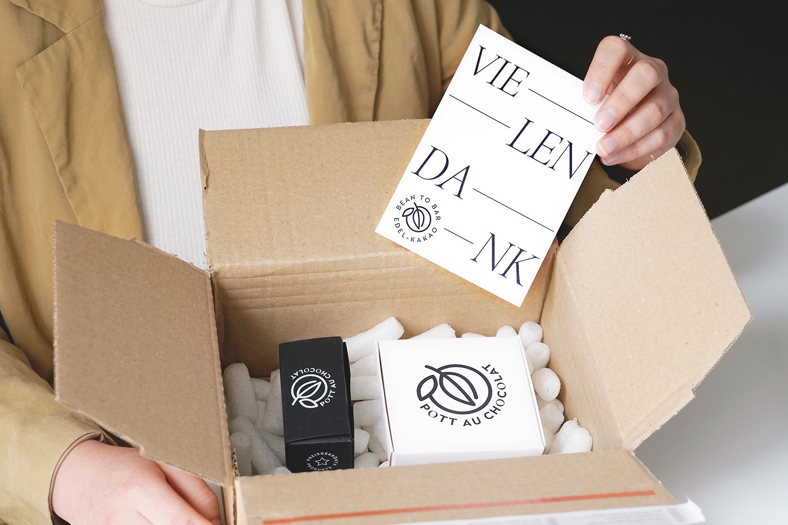 Ein Paket wird gepackt, mit zwei kleinen Pralinenboxen. Eine Karte mit der Aufschrift 'Vielen Dank' wird in das Paket gelegt.