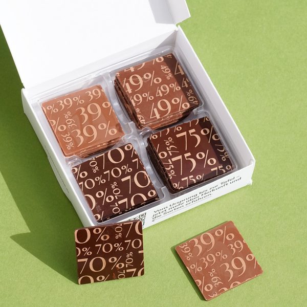 Geöffnete Schachtel mit den Täfelchen sichtbar. 4 verschiedene, die jeweils bedruckt mit farbiger Kakaobutter in Kupfer den Kakaoanteil zeigen.