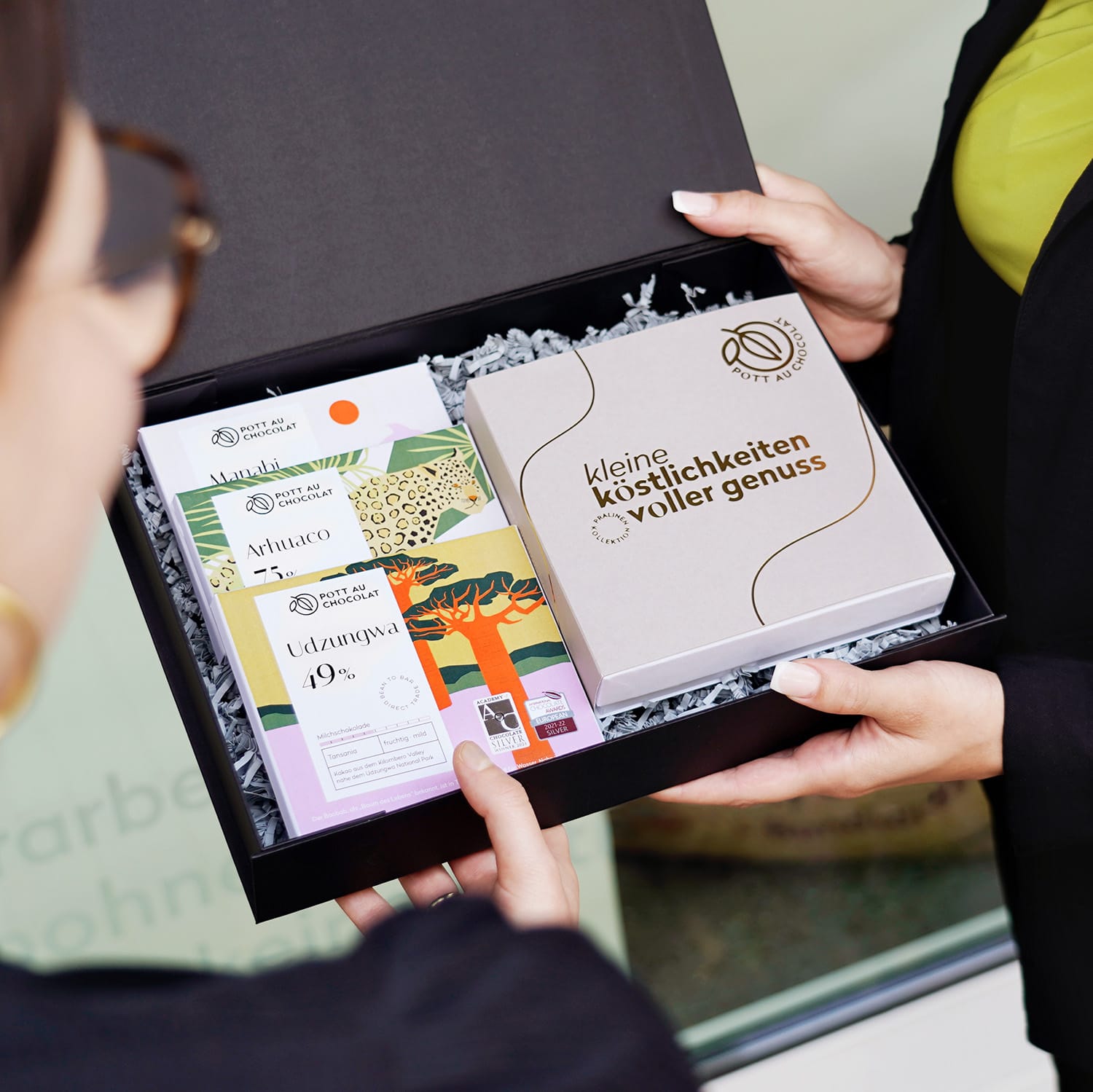 Eine geöffnete Geschenkbox aus schwarzem Karton mit drei Schokoladentafeln und einer Pralinenschachtel wird überreicht.