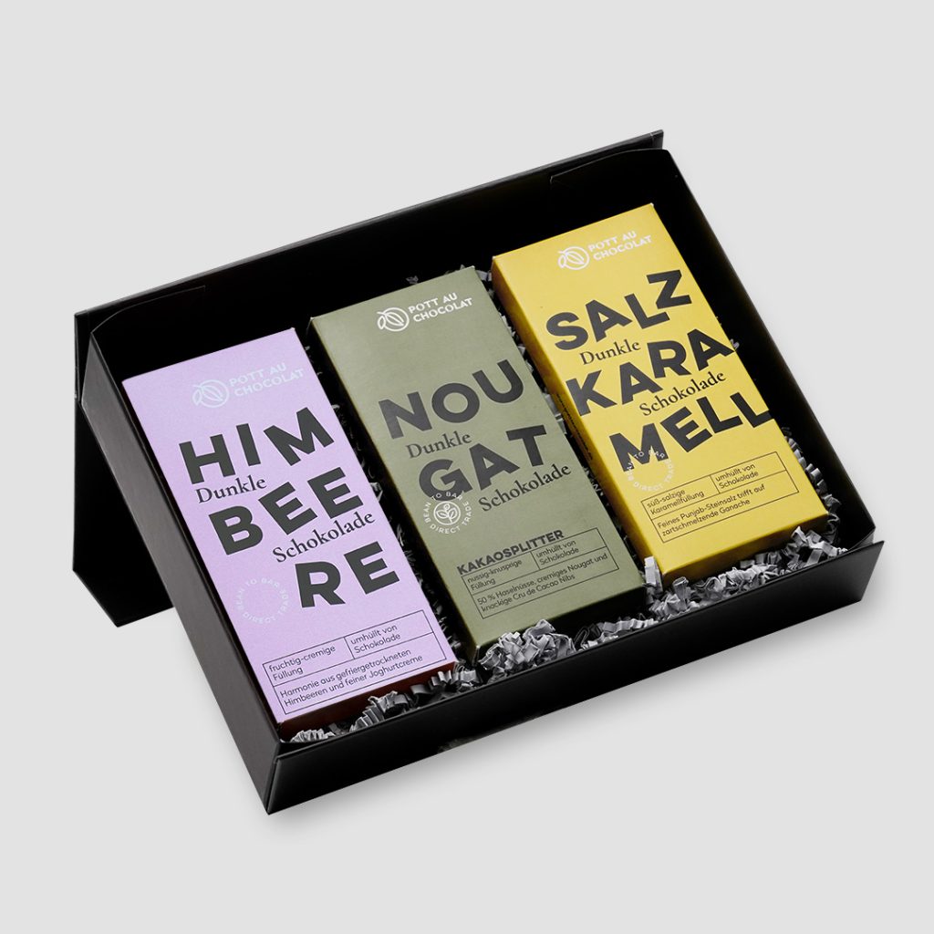 Geöffnete schwarze Geschenkbox mit drei rechteckigen Schokoladentafeln nebeneinander. Die Verpackungen sind einfarbig in rosa, dunkelgrün und gelbgrün mit schwarzer Typografie. Sorten: Himbeere, Nougat Kakaosplitter und Salzkaramell.