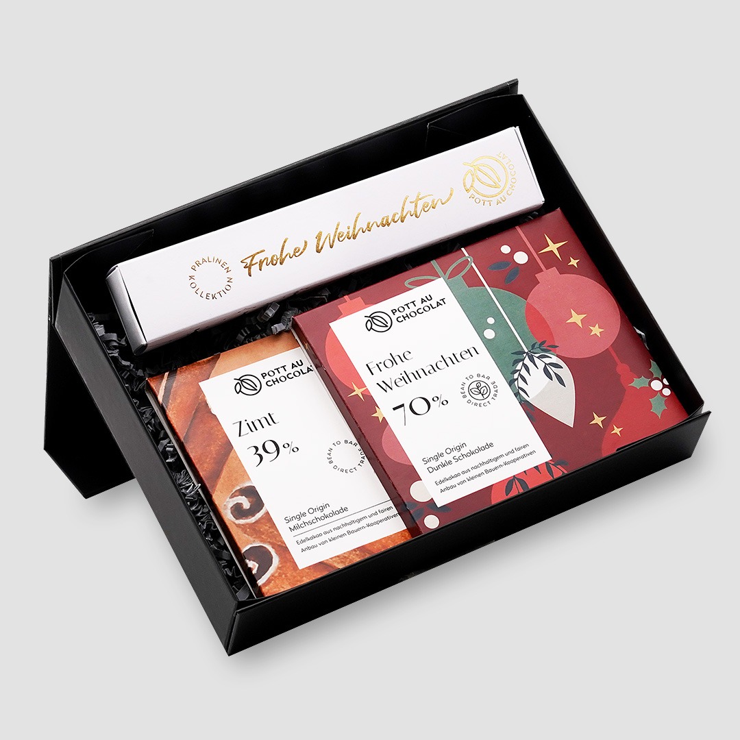 Schwarze Geschenkbox mit einer Stange mit 6 Pralinen mit weißer Banderole mit gold geprägtem 'Frohe Weihnachten'. Zusätzlich zwei Tafeln. Zimt 39% und 'Frohe Weihnachten' 70% im weihnachtlichen Design in Rot, Grün und Weiß.