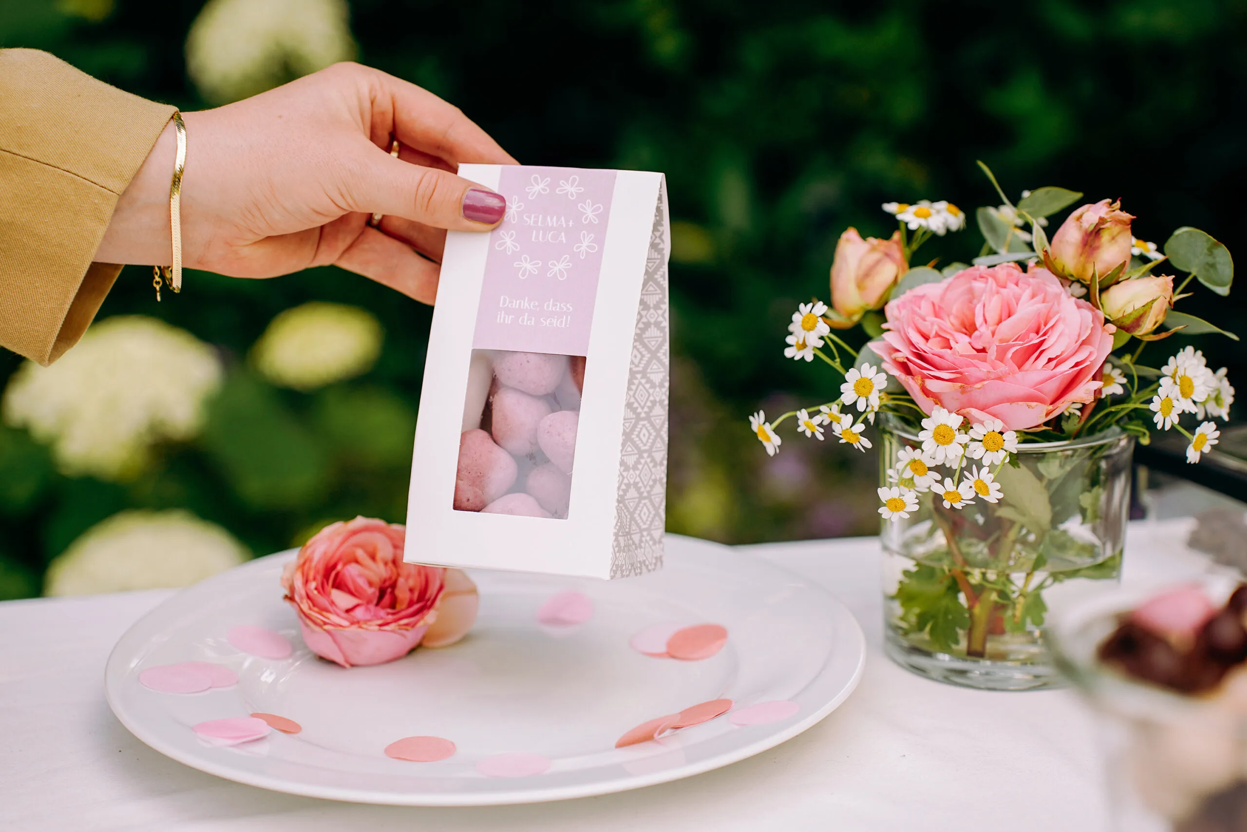 Bild zeigt eine Giebelverpackung mit Fenster von dragierten Himbeeren, die für eine Hochzeitstafel als individuelles Namensschild mit einem eigenen Etikett gestaltet wurde neben einem Frühlingsblumenstrauss