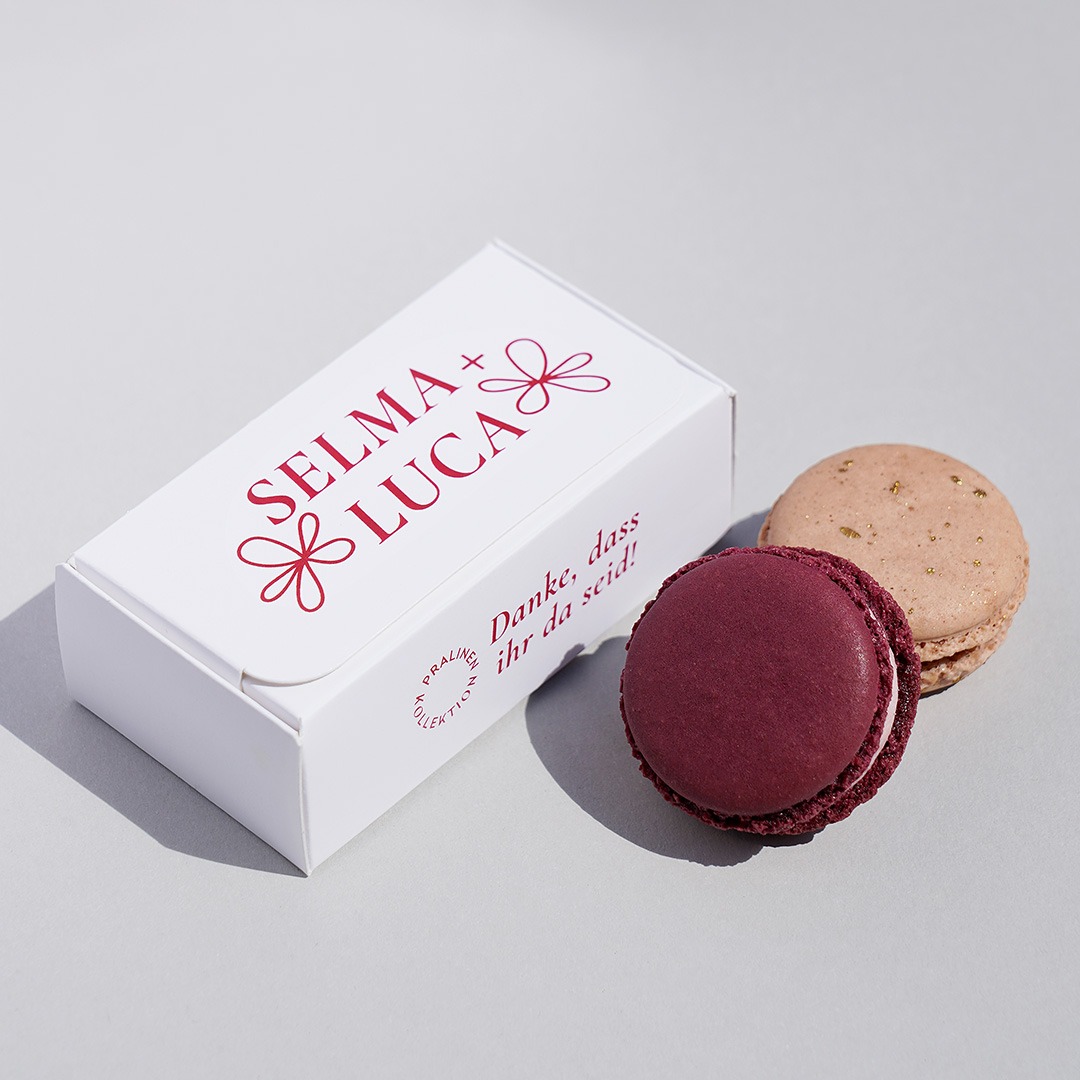 Weiße rechteckige Schachtel, rot bedruckt mit Blumen 'Selma + Luca' und dem Satz: 'Danke, dass ihr da seid!'. Davor liegen zwei Macarons, in Weinrot und Hellbraun mit goldenen Sprenkeln.