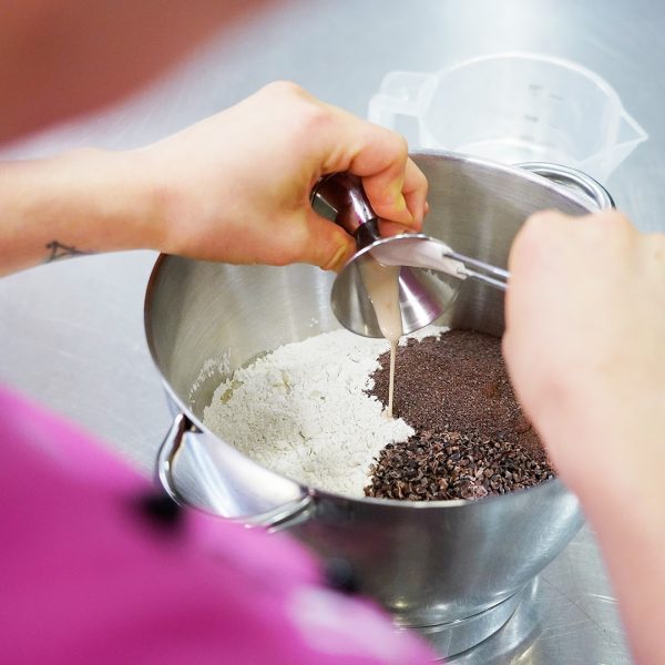 Verschiedene Zutaten für ein Brot werden zusammen in eine Schüssel gegeben. Zum Beispiel Kakaofaser Mehl, Kakao Nibs, Mehl und Hefe.