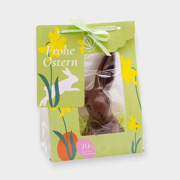 Knickohrhase aus Milchschokolade in grüner Verpackung mit Sichtfenster und Illustrationen: Narzissen, Osterhase, Eier und die Aufschrift 'Frohe Ostern'