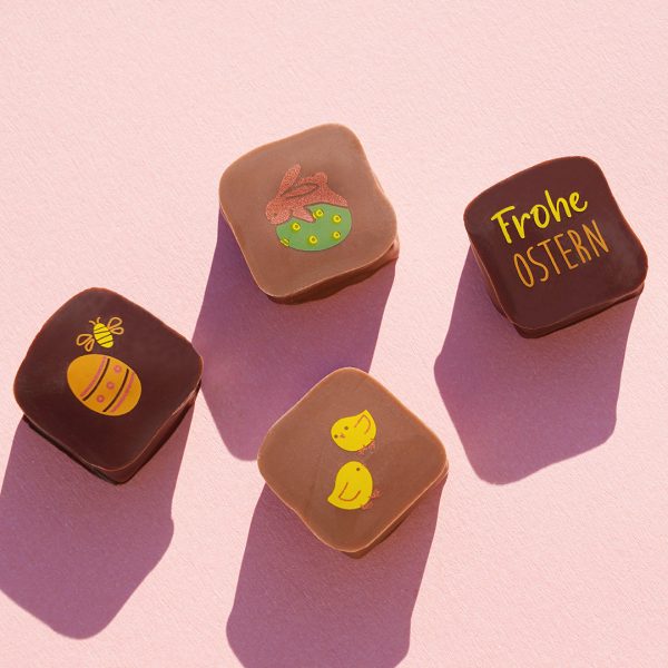 Vier verschiedene Pralinen mit Ostermotiven: ein Häschen auf einem Ei liegend, ein Osterei mit einer Biene, 'Frohe Ostern' und zwei Küken