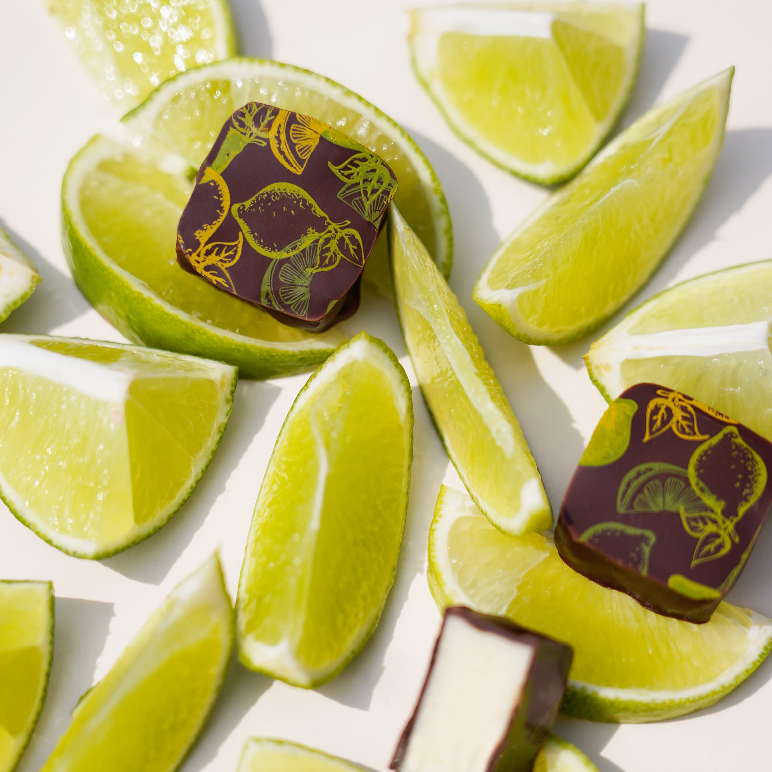 Zwei Caipirinha-Pralinen mit Limettenverzierungen und eine halbierte Praline mit heller Füllung auf einem Hintergrund mit aufgeschnittenen Limetten