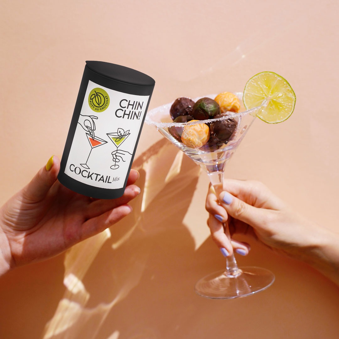 In einer Hand die Cocktail Edition in einer schwarzen Metalldose, in einer anderen Hand ein Cocktailglas mit Pralinen gefüllt. Die Hände stoßen an.