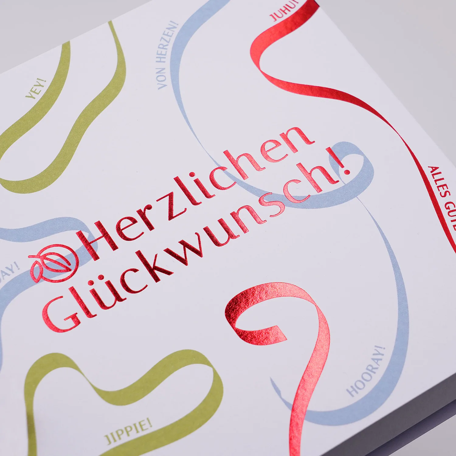 Detail der Pralinen Schachtel 'Herzlichen Glückwunsch' Motiv mit Luftschlangen in rot, blau und grün. Der Hintergrund des Motivs ist weiß, die rote Schrift ist geprägt und schimmert metallisch.