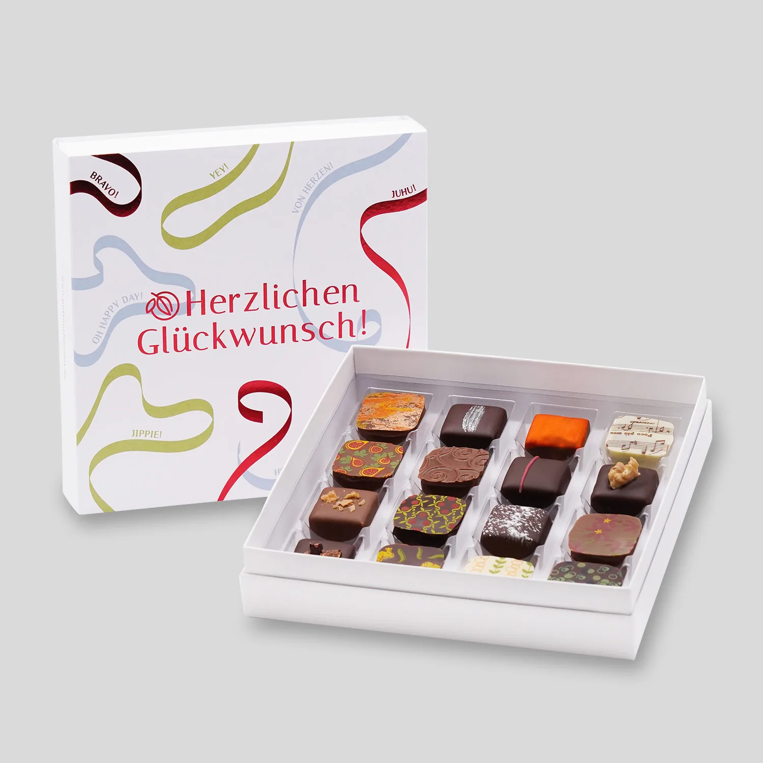 Eine geöffnete Pralinenschachtel steht vor einer geschlossenen mit 'Herzlichen Glückwunsch' Motiv mit Luftschlangen in rot, blau und grün. 16 Pralinen mit verschiedenem bunten Dekor in weißer, dunkler und Milchschokolade.