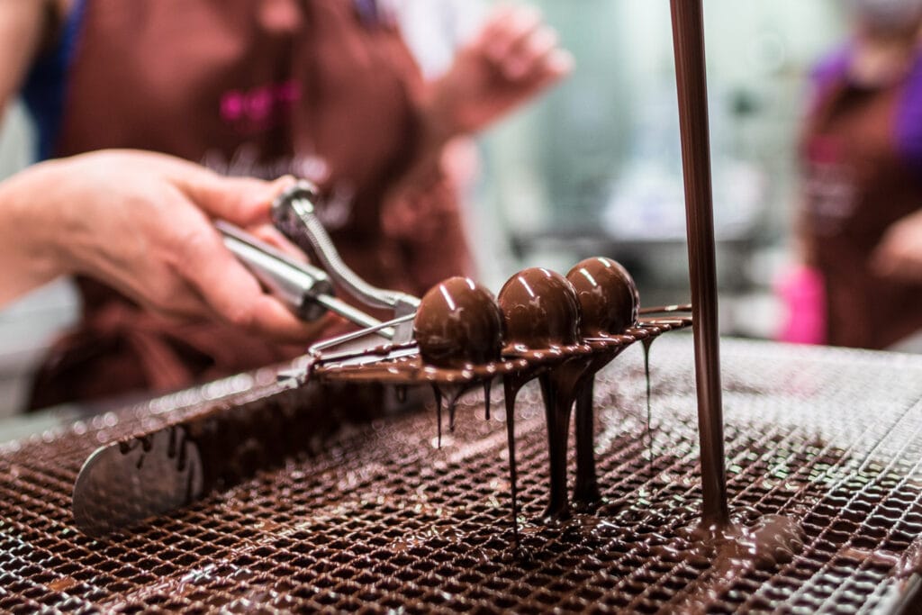 Hand von Kursteilnehmer beim Überziehen von Holkörper Pralinen auf einer breiten Pralinengabel unter einem Schokoladenstrahl