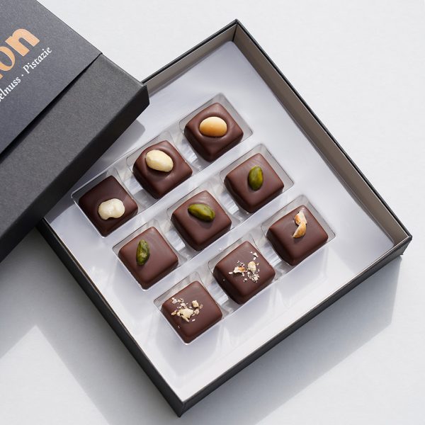 Die geöffnete Schachtel der Nougat Pralinen. Man sieht 9 Pralinen, jeweils 3 dekoriert mit Macadamia, Pistazie und Haselnuss.
