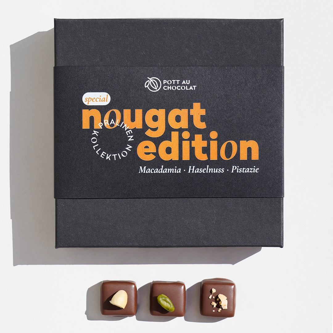 Nougat Pralinen Edition Verpackung. Schwarze Schachtel mit Banderole mit Schrift in orange und weiß. Darunter liegen 3 Pralinen dekoriert mit Macadamia, Pistazie und Haselnuss.