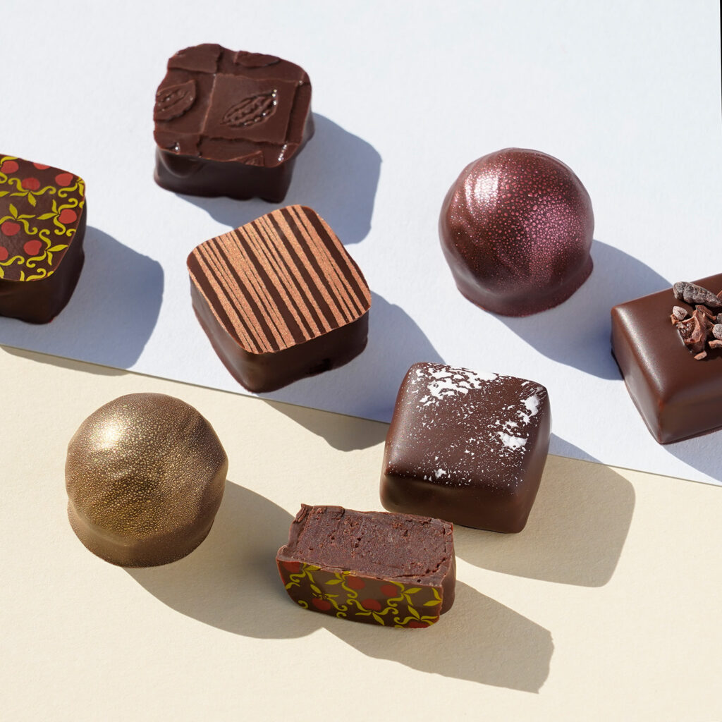 8 verschiedene Pralinen Trüffel, teilweise angeschnitten, dass man die dunkle, cremig feste Ganache sehen kann. Mnache sind rund, manche eckig. Die Pralinen sind dekoriert mit verschiedenen Mustern oder metallisch glänzenden Farben