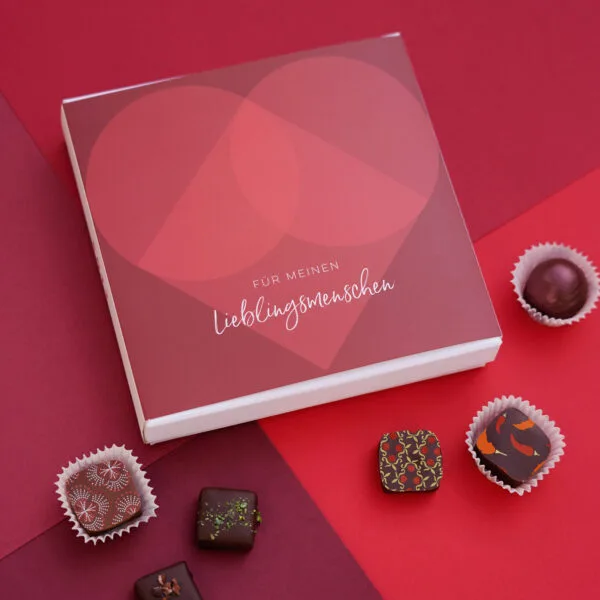 Pralinenbox 'Lieblingsmensch' in weiß mit roter Banderole mit großem roten Herz, davor ein paar bunt dekorierte Pralinen auf rotem Hintergrund.
