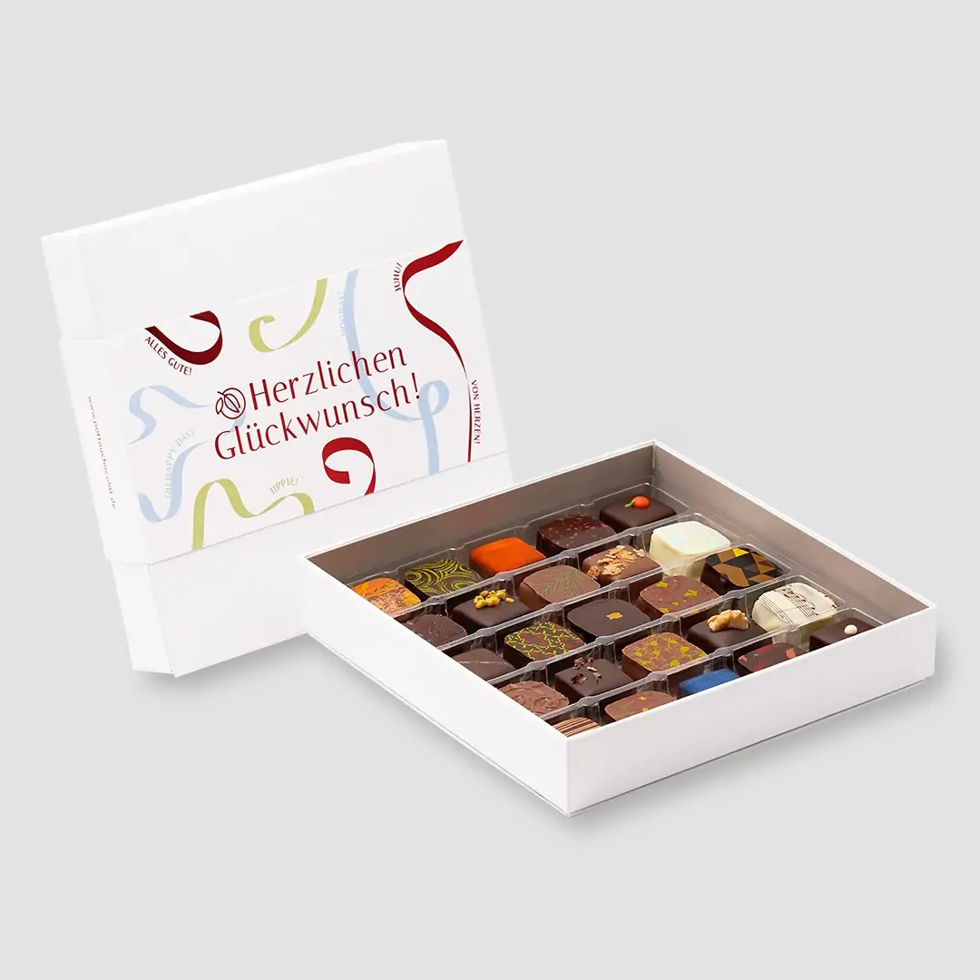 Eine geöffnete Pralinenschachtel mit 25 Pralinen steht vor einer geschlossenen mit 'Herzlichen Glückwunsch' Motiv mit Luftschlangen in rot, blau und grün. 16 Pralinen mit verschiedenem bunten Dekor in weißer, dunkler und Milchschokolade.