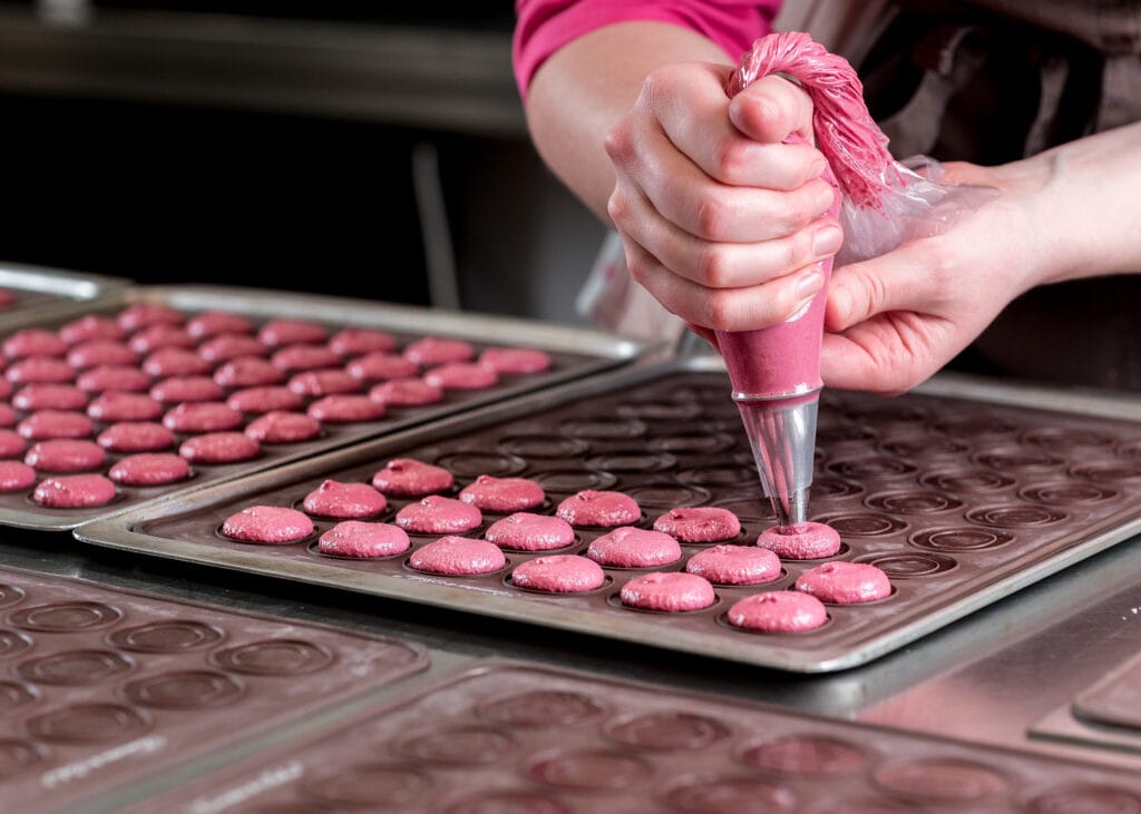 Aus der Produktion: Macarons werden hergestellt. Baiser-Mandel-Masse wird mit einem Spritzbeutel auf eine Macaron-Backmatte gegeben