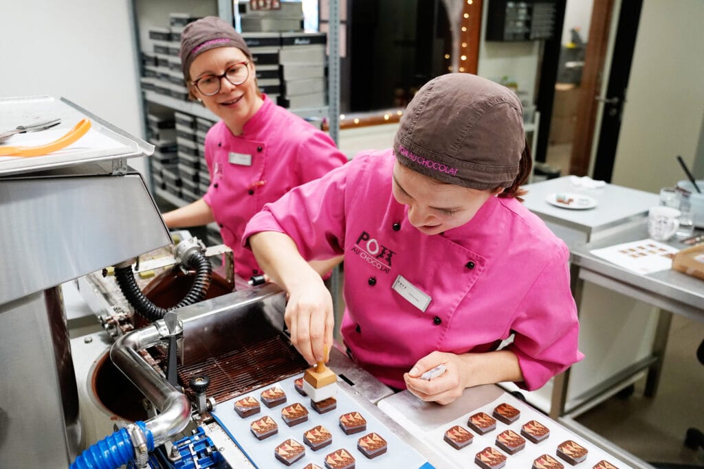 Foto aus der Produktion von Pott au Chocolat: Zwei Mitarbeiter beim Auflegen von Transferfolien aus der Weihnachtskollektion auf große Pralinen