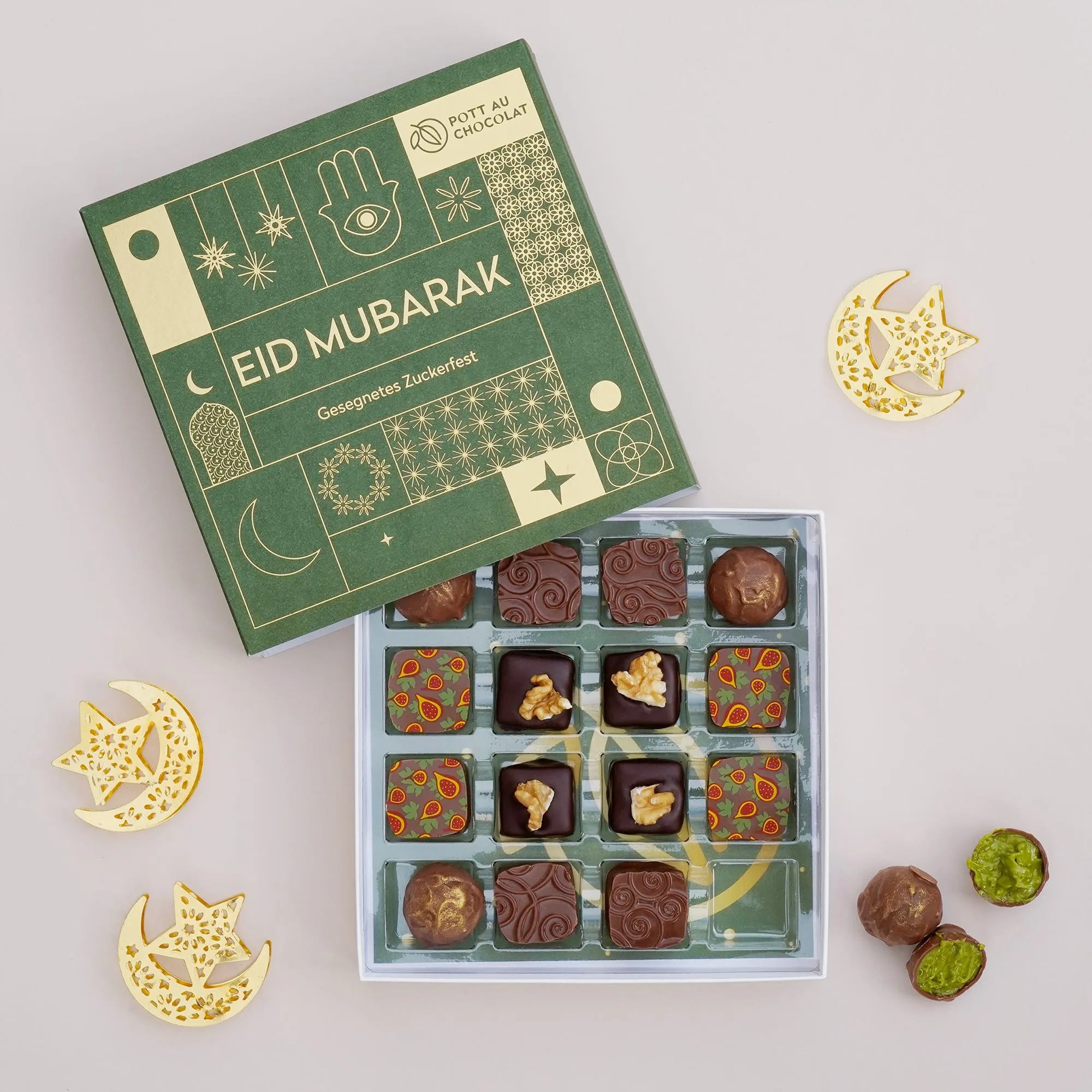 Geöffnete Pralinenschachtel mit 16 Pralinen in dunkler und Milchschokolade, jeweils mit unterschiedlichem Dekor. Auf der Schachtel steht 'Eid Mubarak – Gesegnetes Zuckerfest' mit Illustrationen in goldener Prägung. Goldene Sterne und ein goldener Mond dienen als Dekoration um die Pralinenschachtel herum. Eine Praline 'Dubai' ist aufgeschnitten und liegt ebenfalls neben der Schachtel.