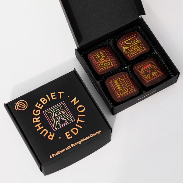 Verpackung der Ruhrgebiet Edition 4er Pralinen. Schwarze Schachtel farbig bedruckt mit Förderturm Motiv. Daneben die geöffnete Verpackung mit 4 Pralinen mit Ruhrgebiets-Motiven in gelb, rot und orange.