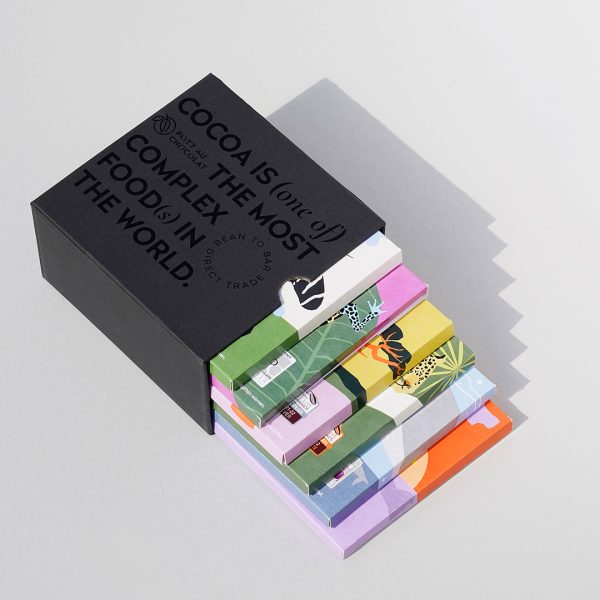 6 bunte Schokoladentafeln in einem schwarzen Schuber aus Pappe, der mit schwarzer Schrift geprägt wurde. Der Text ist englisch und der Inhalt: Kakao ist eines der komplexesten Lebensmittel der Welt. Die Schokoladentafeln sind so aus der Box gezogen, dass man jede etwas sehen kann.